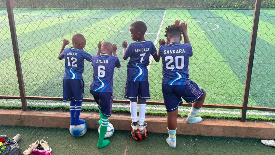 Four young Source of Nile Academy players in blue kit resting on footballs at the pitch side